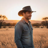 Man wearing a cowboy hat and denim jacket standing in a field at sunset.