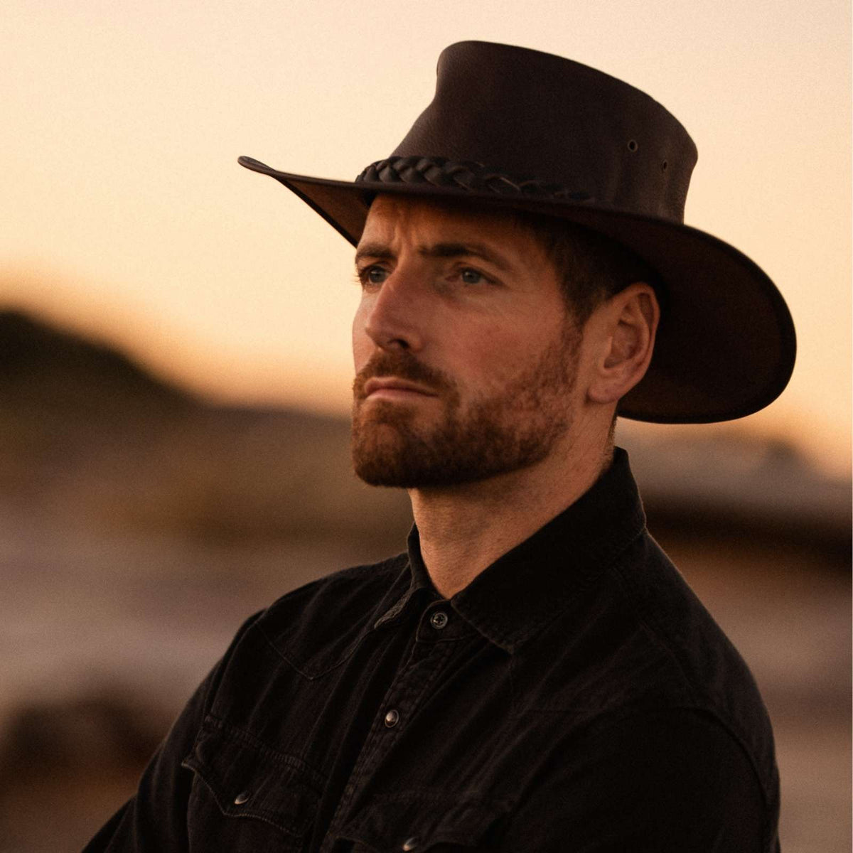 Jacaru 1092 Stockman Hat in dark brown worn by man in black shirt outdoors at sunset.