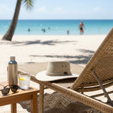 Beach scene with wicker chair, Jacaru Sand Soaker Breeze Hat,glass of water, and bottle on a sandy beach.