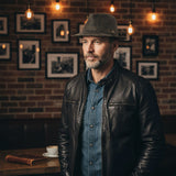 Man wearing a leather jacket and Kangaroo Trilby Hatin a room with framed pictures on the wall.