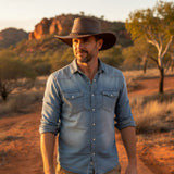 Man wearing a cowboy hat and denim shirt standing in a desert landscape with mountains in the background.