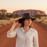 Woman in a white shirt and black hat standing on a dirt road with Uluru in the background