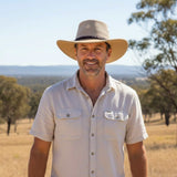 Man wearing a beige hat and shirt standing in a field with trees and mountains in the background