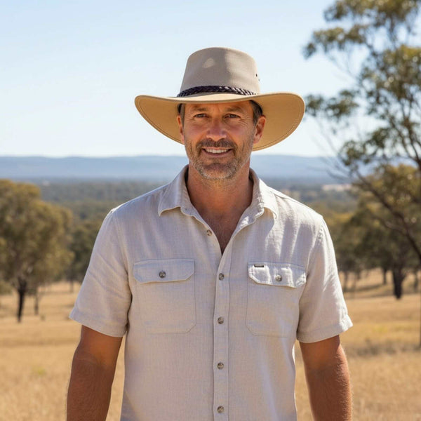Man wearing a beige hat and shirt standing in a field with trees and mountains in the background