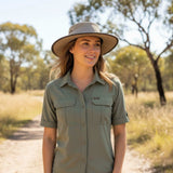 Woman wearing a green shirt and beige Jacaru 104 Bushbreeze Camper Hat in a natural setting with trees and grass.