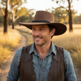 Man wearing a brown cowboy hat and leather vest in a field with trees in the background