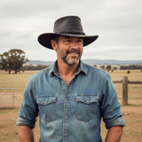 Man wearing a denim shirt and A Black Jacaru 1069 Buffalo Leather Hat standing in a field with trees and a fence in the background.