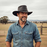 Man wearing a denim shirt and A Black Jacaru 1069 Buffalo Leather Hat standing in a field with trees and a fence in the background.