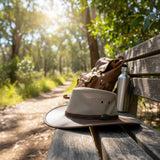 Jacaru Mushroom Safari Hat, backpack, and water bottle on a wooden bench in a forest