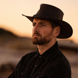 Jacaru 1092 Stockman Hat in dark brown worn by man in black shirt outdoors at sunset.