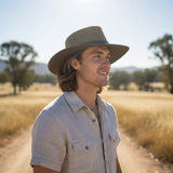 Man wearing a Sunset Breeze Hat in a field with trees and mountains in the background