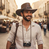 Man wearing a wide-brimmed hat and carrying a camera, standing on a street.
