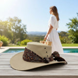 Sand Kangaroo Ladies Breeze Hat with brown band on a wooden surface with a blurred background of a pool and greenery.