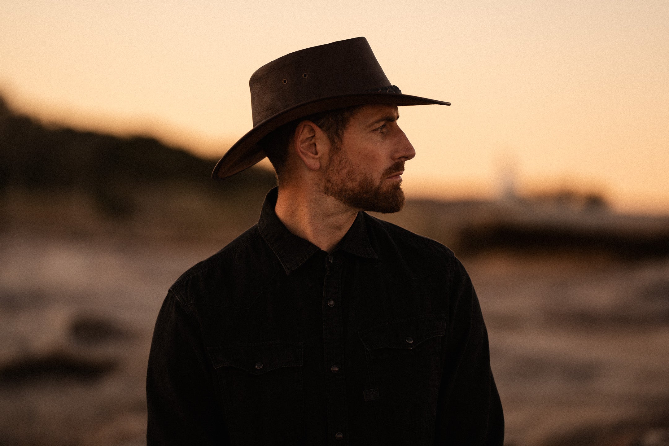 Man wearing a black hat and dark shirt against a sunset background