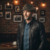 Man wearing a leather jacket and Kangaroo Trilby Hatin a room with framed pictures on the wall.